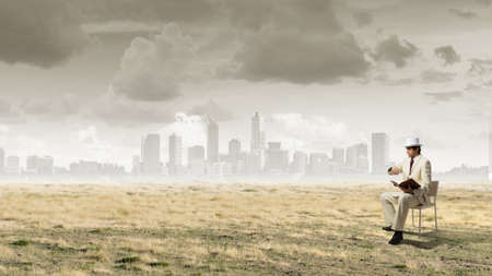 Young businessman sitting in chair with book in hands looking at watch on handの写真素材