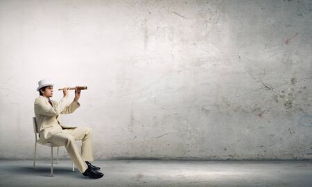 Businessman in white suit and hat sitting in chair and looking in spyglassの写真素材