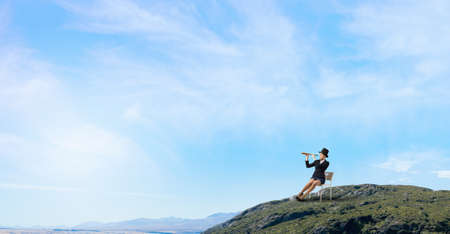 Pretty girl wearing retro hat siting on chair and looking in spyglassの写真素材