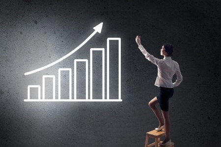 Back view of businesswoman standing on chair and reaching infographs on wallの写真素材