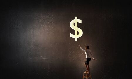 Young attractive businesswoman standing on chair and reaching money bagの写真素材