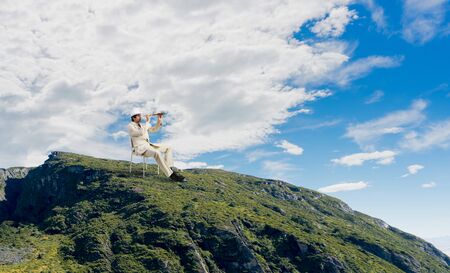 Businessman in white suit and hat sitting in chair and looking in spyglass