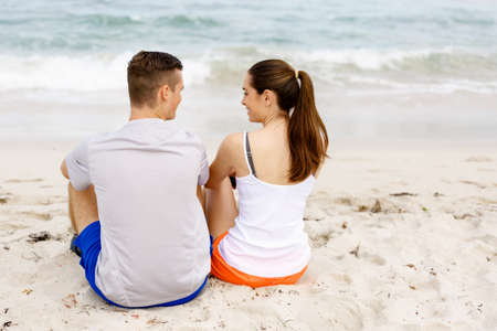 Young couple looking at each other while sitting together on beach in sports wearの写真素材