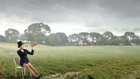 Pretty girl wearing retro hat siting on chair and looking in spyglassの写真素材