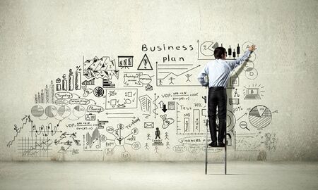 Back view of businessman standing on ladder and drawing sketches on wallの写真素材