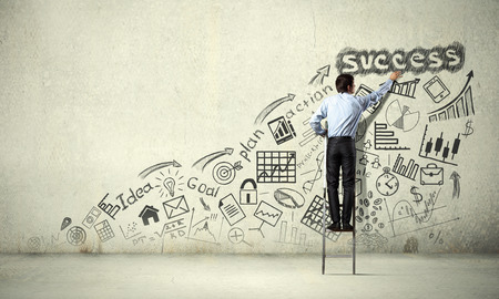 Back view of businessman standing on ladder and drawing sketches on wallの写真素材