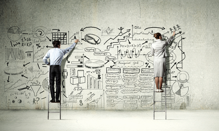 Back view of businessman and businesswoman standing on ladder and drawing sketches on wallの写真素材