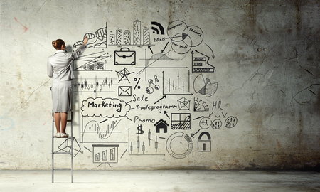Back view of businesswoman standing on ladder and drawing sketches on wallの写真素材