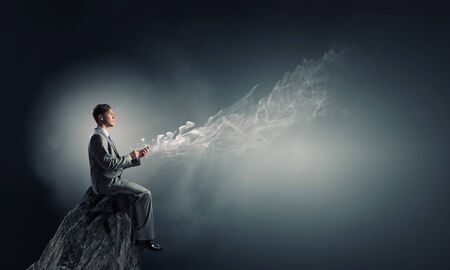 Young businessman sitting on top of rock with mobile phone in handの写真素材