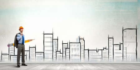 Young male engineer in hard hat with big pencil and buildings sketches at backgroundの写真素材