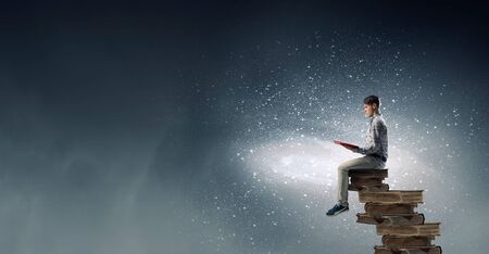 Young man sitting on pile of old books with one in handsの写真素材