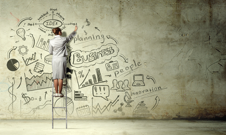 Back view of businesswoman standing on ladder and drawing her great plan on wallの写真素材