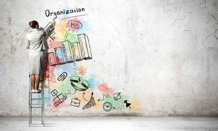 Back view of businesswoman standing on ladder and drawing sketches on wallの写真素材
