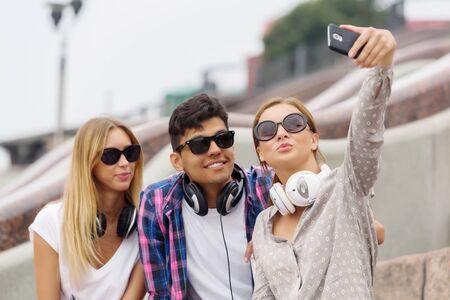 Three young happy people sitting outdoors and making selfie