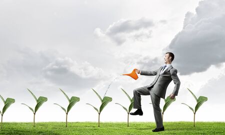 Young cheerful businessman watering green sprout with canの写真素材