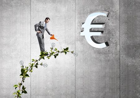 Young cheerful businessman watering green euro sprout with canの写真素材