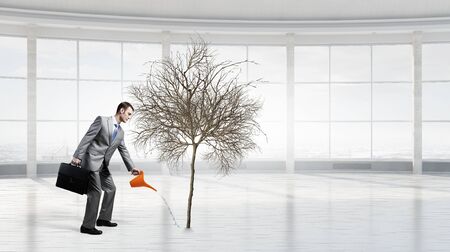 Young cheerful businessman watering dry tree with canの写真素材
