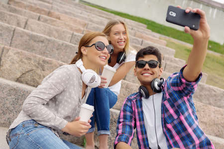 Three young happy people sitting outdoors and making selfie