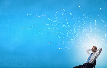 Young businessman sitting in chair with ideas coming out of his headの写真素材