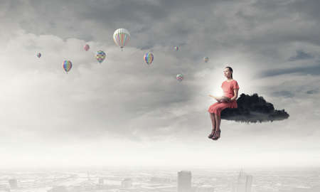 Young woman in dress with old book in hand sitting on cloudの写真素材