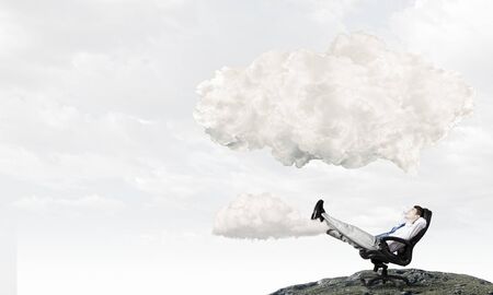 Relaxed businessman with hands behind his head sitting in chair and thought cloud aboveの写真素材