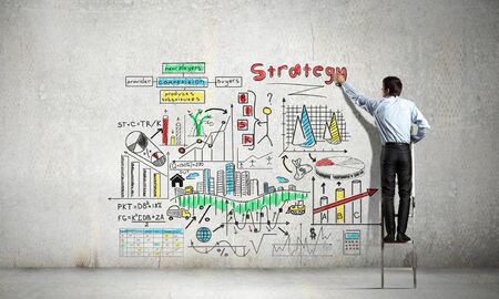 Back view of businessman standing on ladder and drawing sketches on wallの写真素材