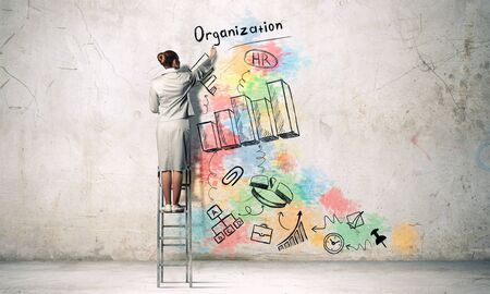 Businesswoman standing with back on ladder and drawing ideas on wallの写真素材