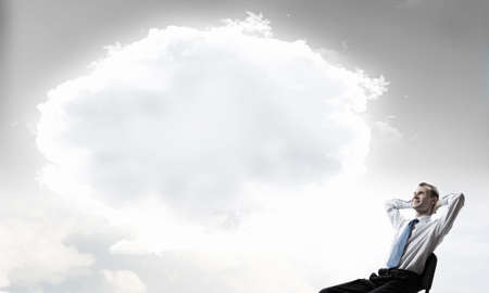 Relaxed businessman with hands behind his head sitting in chair and thought cloud aboveの写真素材
