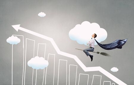 Young businessman wearing cape flying on broom high in skyの写真素材