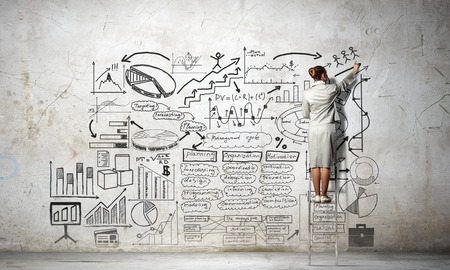 Rear view of businesswoman standing on ladder and drawing business sketch on wallの写真素材