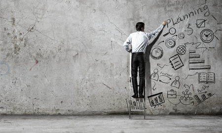 Businessman standing with back on ladder and drawing ideas on wallの写真素材