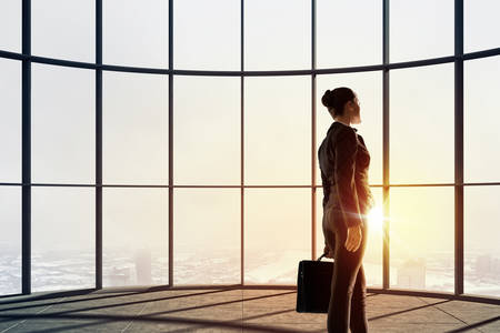 Back view of businesswoman in modern office facing sunriseの写真素材