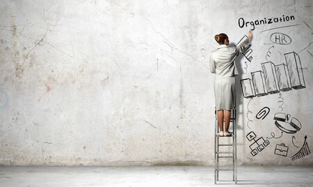 Businesswoman standing with back on ladder and drawing ideas on wallの写真素材