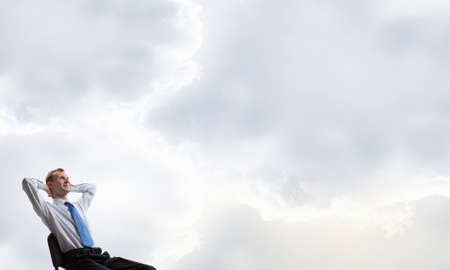 Relaxed businessman with hands behind his head sitting in chair and thought cloud aboveの写真素材