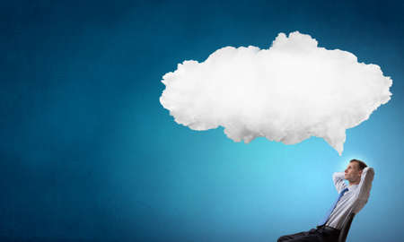 Relaxed businessman with hands behind his head sitting in chair and thought cloud aboveの写真素材
