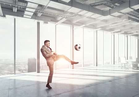 Businessman in suit in modern office jumping to hit soccer ballの写真素材