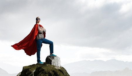 Young confident woman in red cape and mask on rock topの写真素材