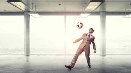 Businessman in suit in modern office jumping to hit soccer ballの写真素材