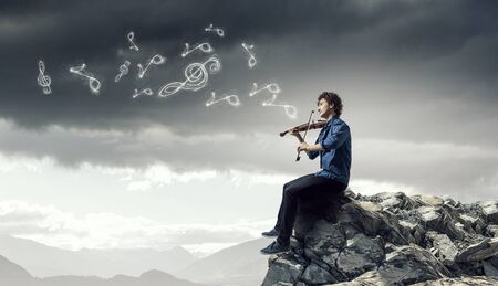 Young handsome man sitting on rock edge and playing violin