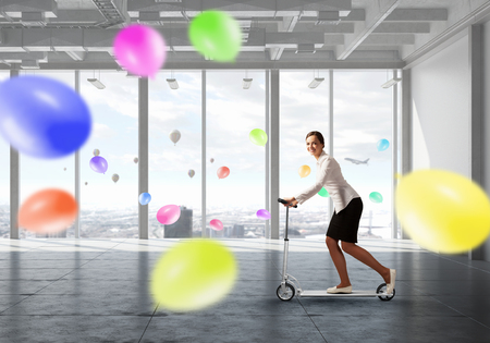 Young cheerful businesswoman in suit riding scooter in officeの写真素材