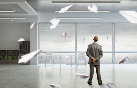 Thoughtful businessman standing with back in empty officeの写真素材