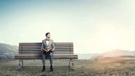 Young businessman wearing red bow tie sitting on bench with book in handsの写真素材