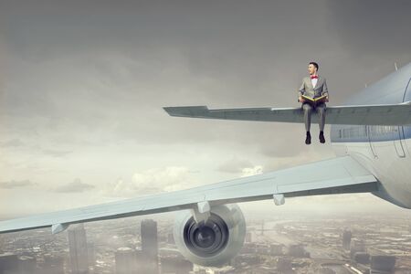 Young businessman sitting on wing of airplane and reading bookの写真素材