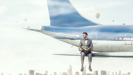 Young businessman sitting on wing of airplane and reading bookの写真素材