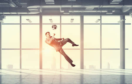 Businessman in suit in modern office jumping to hit soccer ballの写真素材