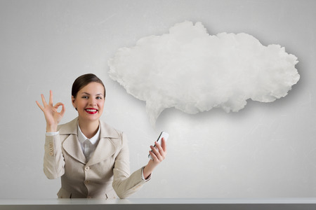 Young pretty businesswoman sitting at table with mobile phone in hand showing ok gestureの写真素材