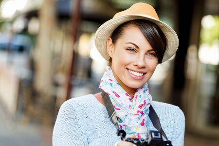Outdoor summer smiling lifestyle portrait of pretty young woman having fun in the city with cameraの写真素材