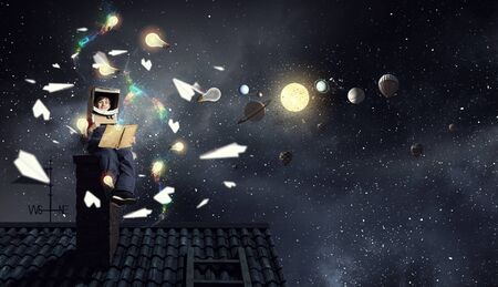 Funny boy in carton helmet reading book and dreaming he is astronaut