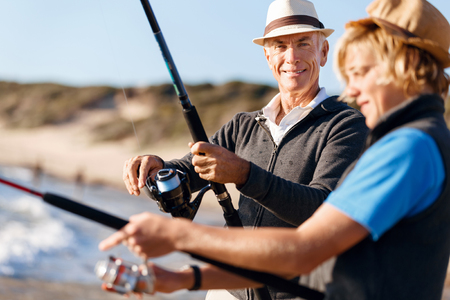 Senior man fishing with his teenage grandson at seasideの写真素材