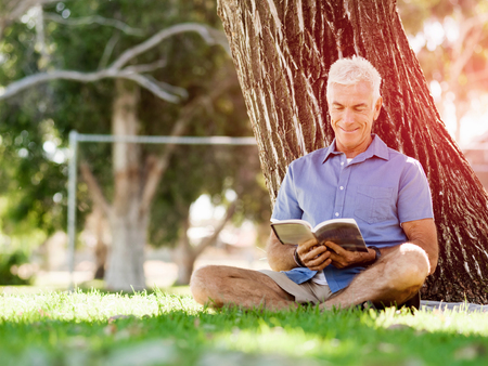Handsome senior sitting in park and reading bookの写真素材